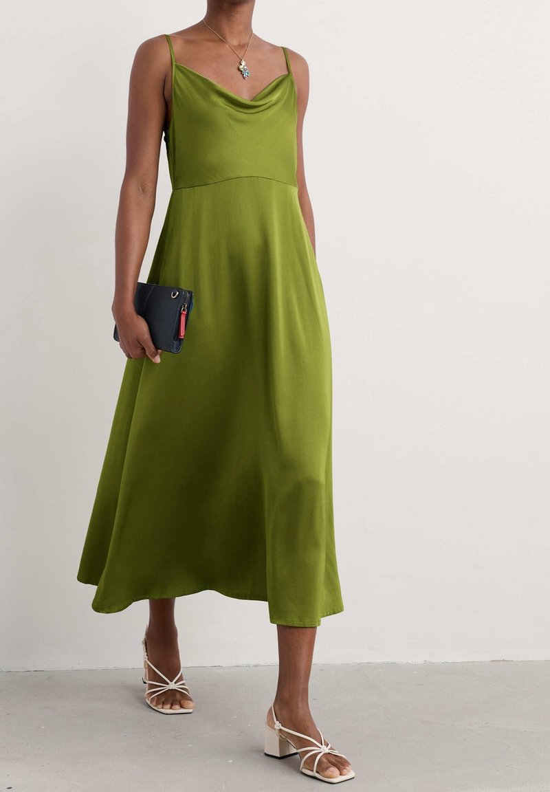 Robe en satin vert avec un décolleté drapé et de fines bretelles. Associée à une pochette noire et des sandales blanches à talons blocs avec des lanières. Texture lisse.