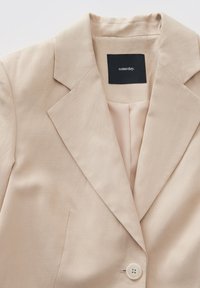 Blazer beige con un design strutturato, dotato di chiusura con un solo bottone, rever a taglio e un tessuto liscio e texture. Etichetta visibile all'interno.