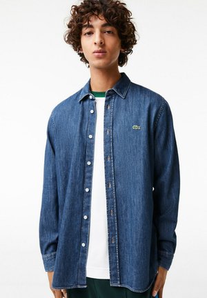 Denim shirt in dark blue with a button-down design, featuring long sleeves and a small green logo on the chest. Smooth texture with a relaxed fit.