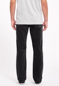 Pantalons en velours côtelé noir avec une coupe droite, tissu texturé, poches arrière, et portés avec des baskets basses. Tatouage visible sur le poignet.