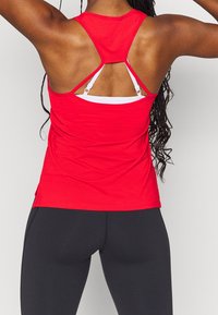 Rode sportieve tanktop met een racerback ontwerp en een mesh textuur. Gelaagd met een witte sportbeha eronder. Gecombineerd met zwarte leggings.