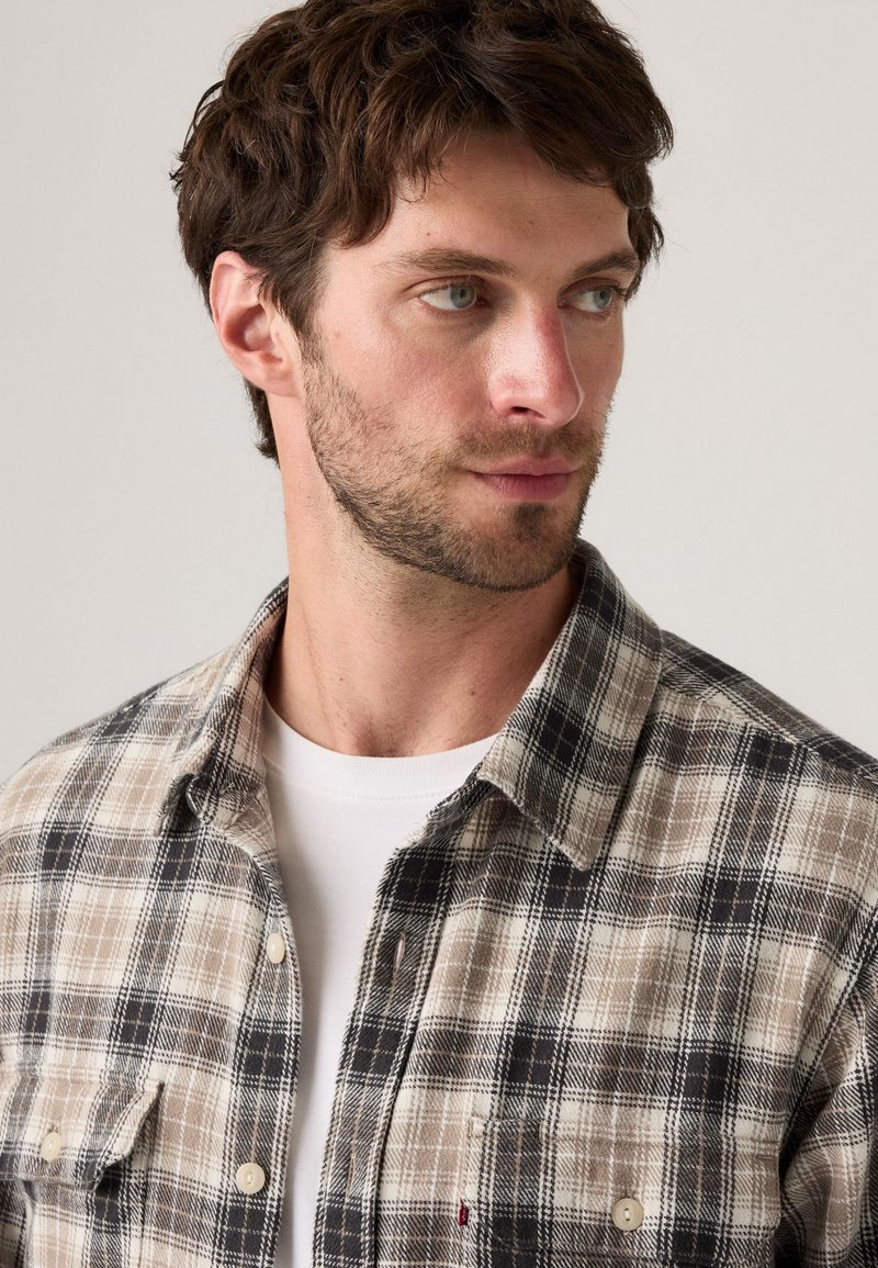 Plaid flannel shirt in beige, black, and white; button closure; two chest pockets; soft texture; collar design; worn over a white t-shirt.