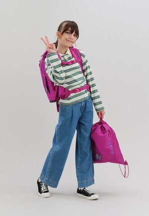 Mädchen trägt gestreiftes Hemd und Jeans, hat einen pinkfarbenen Rucksack und eine Tasche dabei, macht mit der rechten Hand ein Peace-Zeichen.