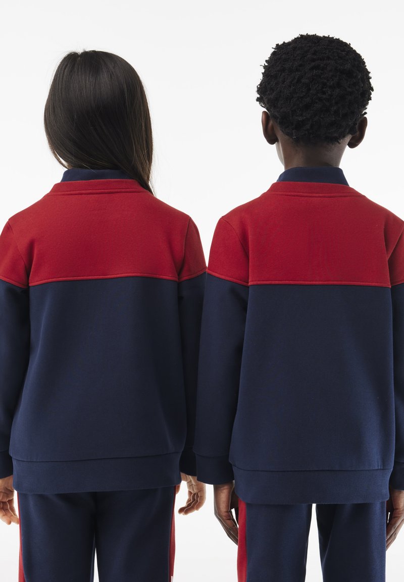 Sweatshirts bleu marine et rouge à blocs de couleur avec un col, présentant des manches longues et un tissu texturé. La vue de dos montre une coupe décontractée.