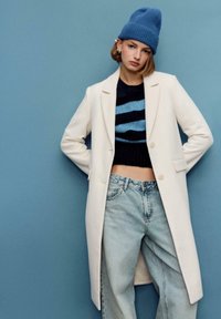 Cappotto bianco, maglione corto a righe blu navy e azzurro, jeans larghi azzurri, berretto blu, modella in piedi davanti a uno sfondo blu uniforme.