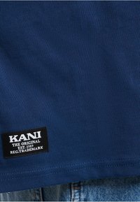 Marineblå ribbet T-skjorte med en svart etikett som har "KANI THE ORIGINAL EST. 1989" i hvit tekst nederst til venstre.