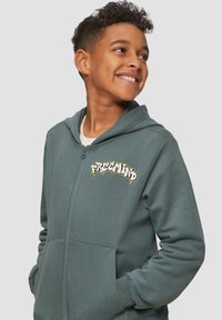 Dunkelgrüne Zip-Hoodie aus weichem Material mit einer Vordertasche und dem „FREEMIND“-Logo in Weiß und Gelb auf der Brust.
