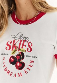 Bílá tričko s červeným výstřihem a lemy rukávů. Obsahuje grafiku třešně a text: "Třešňová nebe, sny o oči," s nápisem "100% organické."