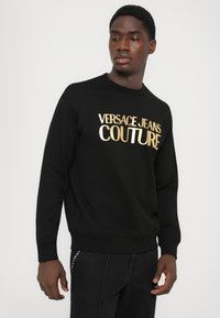 Črna felpa z zlatim napisom "VERSACE JEANS COUTURE". Narejena iz mehkega materiala, z okroglim izrezom in rebrastimi manšetami.