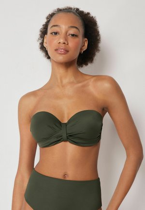 Junge Frau mit lockigem Haar, die ein olivgrünes, trägerloses Bikinioberteil und passende, hoch taillierte Höschen vor einfarbigem Hintergrund trägt.