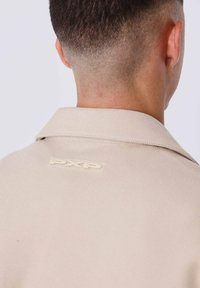 Giacca beige in tessuto testurizzato, con colletto a punte e un'etichetta logo "PXP" visibile sul retro.