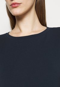 Zbliżenie na kobietę w ciemnoniebieskiej koszulce z okrąg�łym dekoltem, pokazującą szyję, dolną część twarzy i proste brązowe włosy.
