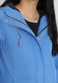 Mujer vestida con una chaqueta con capucha de color azul claro, con camisetas de capas negras y grises debajo, mostrando detalles de frente con cremallera y botones.