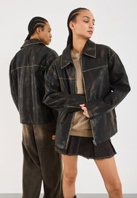 Veste en simili cuir noir avec une finition brillante, présentant un large col et des lignes de couture contrastantes, portée sur des vêtements beiges et sombres.