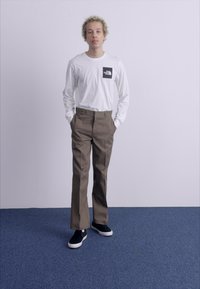Long-sleeve white shirt with a black logo pocket, paired with brown trousers and black sneakers with white soles. Model stands with hands in pockets.