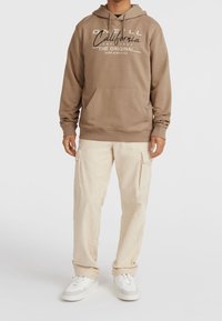 Homme portant un sweat à capuche beige clair avec le texte "O'Neill California Santa Cruz", un pantalon cargo beige et des baskets blanches, debout sur un fond blanc.