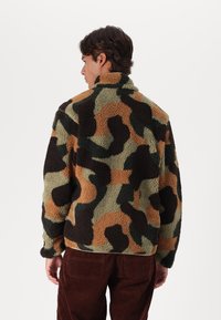 Giacca in pile con motivo camouflage caratterizzata da tonalità di verde, marrone e beige. Tessuto testurizzato con colletto alto e vestibilità morbida.