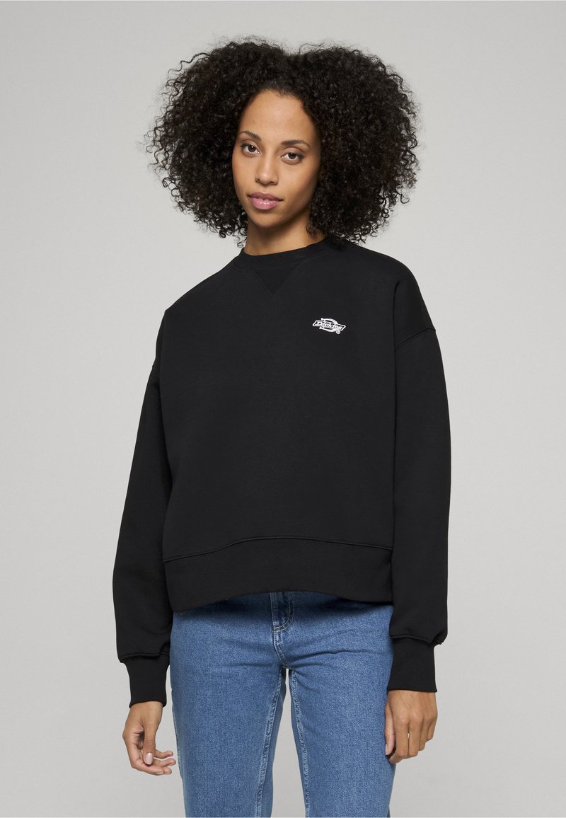 Vrouw met krullend haar die een zwart sweatshirt draagt met een klein wit logo op de borst en blauwe spijkerbroek, staand tegen een effen lichte achtergrond.