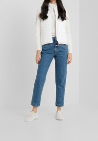 White, quilted sleeveless vest over a cream sweater, paired with mid-rise blue jeans and light-colored sneakers. Simple, casual outfit.