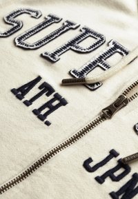 Sudadera cremada con cremallera que presenta el texto bordado en navy "SUPER" y "ATH", con cremallera de metal y textura de algodón.