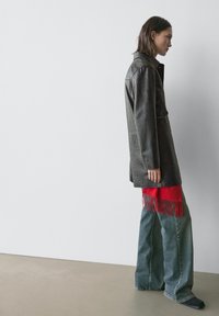 Veste en cuir noir à finition texturée, coupe oversize, associée à un jean large et un foulard rouge à franges. Silhouette simple et moderne.