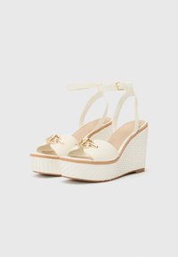 Sandales à talons blancs avec une plateforme tissée texturée, un design à bout ouvert, une lanière ajustable au niveau de la cheville et une embellissement en ton doré à l'avant.
