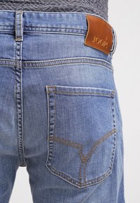 Jean en denim bleu clair avec une étiquette en cuir marron portant la mention "JOOP!" à l'arrière. Présente un design de poche classique avec des coutures contrastantes.