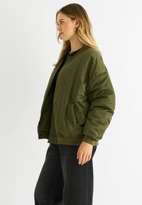 Olive green bomber jacket with a smooth, shiny finish, ribbed collar and cuffs, snap-button closure, and side pockets. Worn over a black shirt.