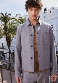 Grå vævet jakke med krave og knapper, der har to forlommer; båret over en farverig skjorte med paisley mønster. Palmetræer i baggrunden.