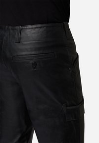 Zwarte leren broek met een gladde textuur, uitgerust met een achterzak met knoopsluiting en zijzakken met stoffen accenten.