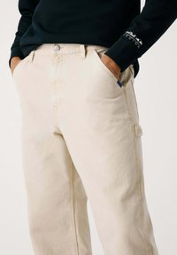 Beige Baumwoll-Cargo-Hosen verfügen über Seitentaschen, einen Frontknopfverschluss und kontrastierende Nähte, mit einer glatten Textur und regulärer Passform.