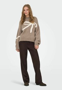 Pull en tricot marron avec un motif abstrait blanc, col et poignets côtelés, associé à un pantalon large marron foncé et des chaussures foncées.