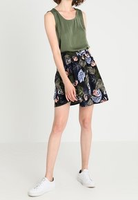 Top a manica lunga verde oliva abbinato a una gonna nera con un motivo di foglie tropicali colorate. Indossato con sneakers bianche.