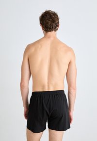Juodi medvilniniai apatiniai šortai su elastinga juosta, eksponuojami ant nuogo torsos, pabrėžiant audinio lygų tekstūrą ir paprastą dizainą.