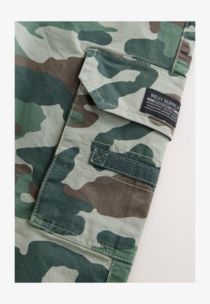 Camouflagemønstret stof i grøn, brun og beige. Har en klaplomme med et mærke og kontrasterende syninger langs kanterne. Holdbart bomuldsmateriale.