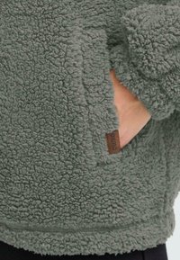 Grüner Fleece-Pullover mit einer weichen, strukturierten Oberfläche, ausgestattet mit einer Seitentasche und einem kleinen braunen Logo-Tag. Gemütliches und lässiges Design.