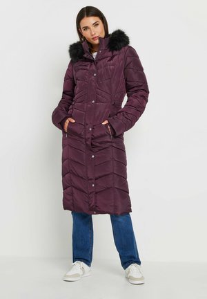 Manteau doudoune long, violet foncé avec un motif en chevrons, bordure en fausse fourrure sur la capuche, fermeture à pression et poches latérales. Porté sur un jean bleu.