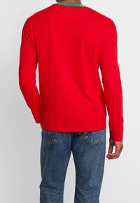 Camisa roja de manga larga de algodón, con cuello redondo y detalle verde. La parte trasera presenta una textura suave y un diseño sencillo.