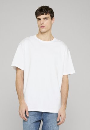 Ung mand med kort krøllet hår iført en ensfarvet hvid oversized t-shirt og blå jeans, stående foran en grå baggrund.