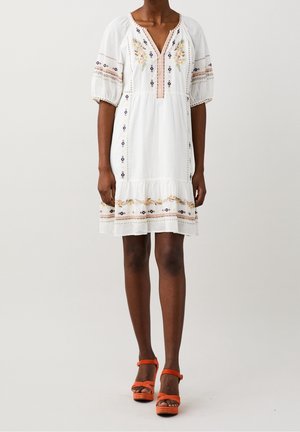 Vrouw met een witte jurk tot op de knie met bloemrijke borduursels en pofmouwen, gecombineerd met oranje hoge hakken.