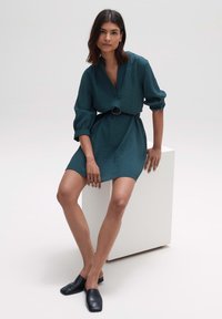 Tee-farbenes Minikleid aus strukturiertem Stoff, mit V-Ausschnitt und langen, gerafften Ärmeln, tailliert mit einem schwarzen Gürtel. Schwarze Slipper.