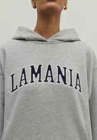 Šedá mikina s kapucí, na které je vyšívané slovo "LAMANIA" námořnickou modří přes hrudník. Měkká textura, volný střih, klokaní kapsa.