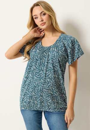 Frau mit langen blonden Haaren, die ein blau-weißes kurzärmeliges Oberteil mit Blattmuster und blaue Jeans trägt, posiert mit einer Hand in der Nähe ihres Halses.