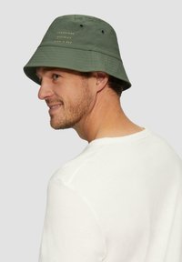 Grüner Bucket Hat aus Stoff, mit Ösen und goldener Schrift: "LAKESHORE DISTRICT FISH-O-REE." Flaches, breites Design.