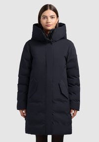 Mornarsko plavi pufasti kaput s visokim ovratnikom, prednjim zatvaračem i dva bočna džepa; glatka tkanina s blago sjajnom završnom obradom.