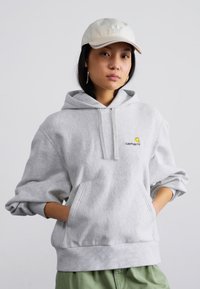 Gray hoodie with a front pocket, drawstrings, and a small yellow and black logo. Worn with a light-colored cap and green pants.
