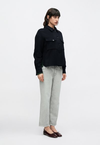 Μπλε πλεκτό σακάκι με δύο τσέπες στο στήθος, κλείσιμο με κουμπιά και γιακά, συνδυασμένο με ανοιχτό γκρι τζιν κανονικής γραμμής και γυαλιστερά μπορντό φλατ παπούτσια.