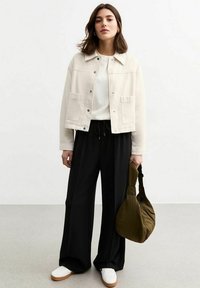 Veste en denim beige clair avec deux poches avant, associée à une chemise blanche et un pantalon noir à jambes larges. Le mannequin tient un sac vert.
