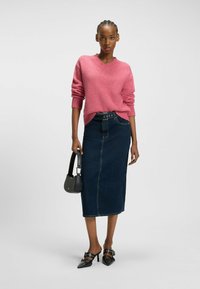 Rosa gestrickter Pullover mit langen Ärmeln, kombiniert mit einem dunkelblauen Denim-Midikleid. Eine schwarze Handtasche und spitz zulaufende schwarze Absatzschuhe runden das Outfit ab.