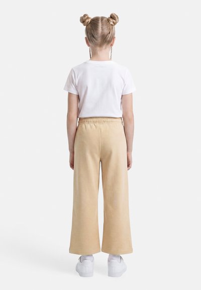 Παιδί που στέκεται με την πλάτη στραμμένη, φοράει λευκό κοντομάνικο πουκάμισο, μπεζ φαρδιά παντελόνια, λευκά αθλητικά παπούτσια και τα μαλλιά του είναι χτενισμένα σε δύο κότσους.